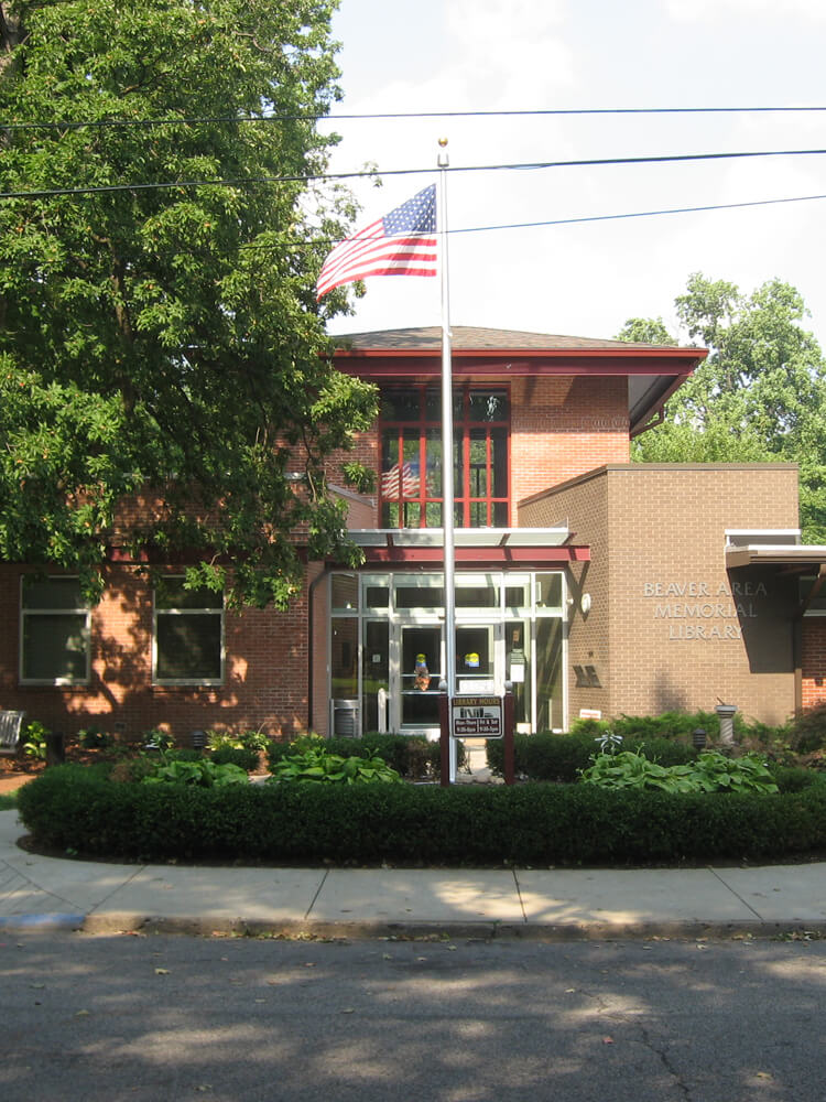 Exterior shot of the Beaver Area Memorial Library building with a prominent American flag and surrounding green hedges .