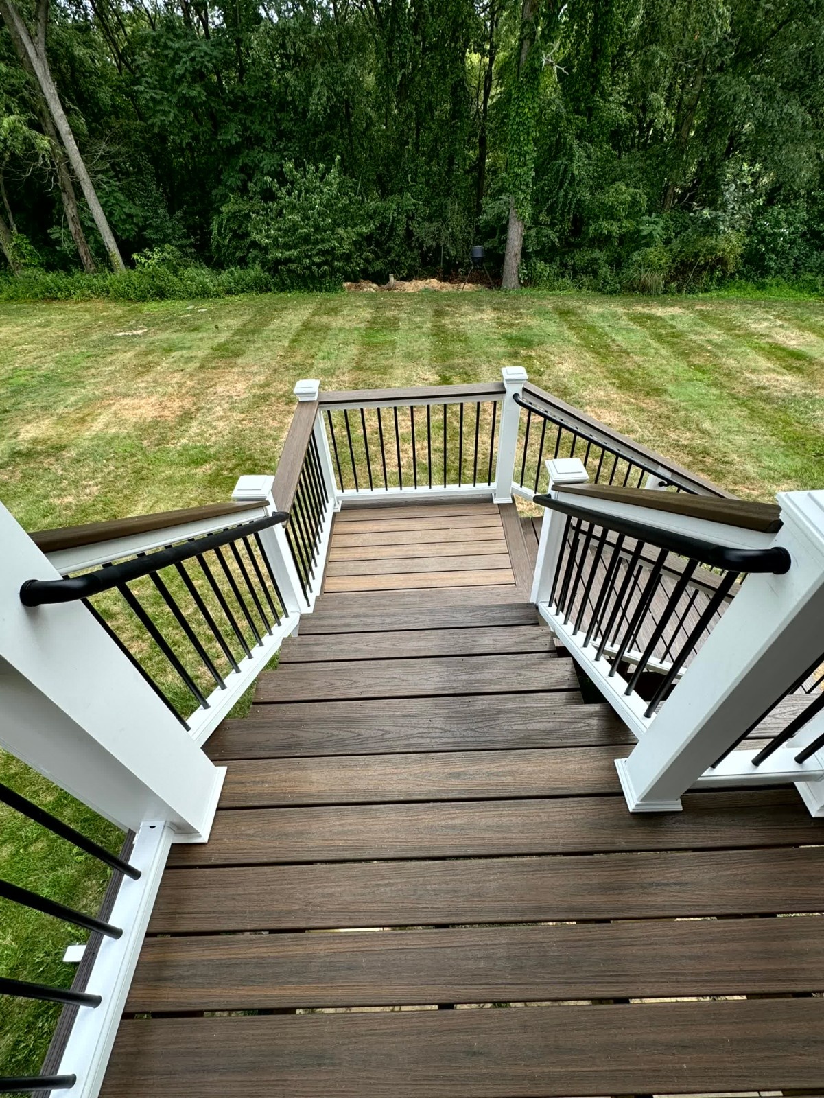 Deck Builder in Monaca, PA: A top-down view of newly installed composite deck stairs featuring dark brown decking, white posts, and contrasting black aluminum railings leading down to a green lawn and wooded area.