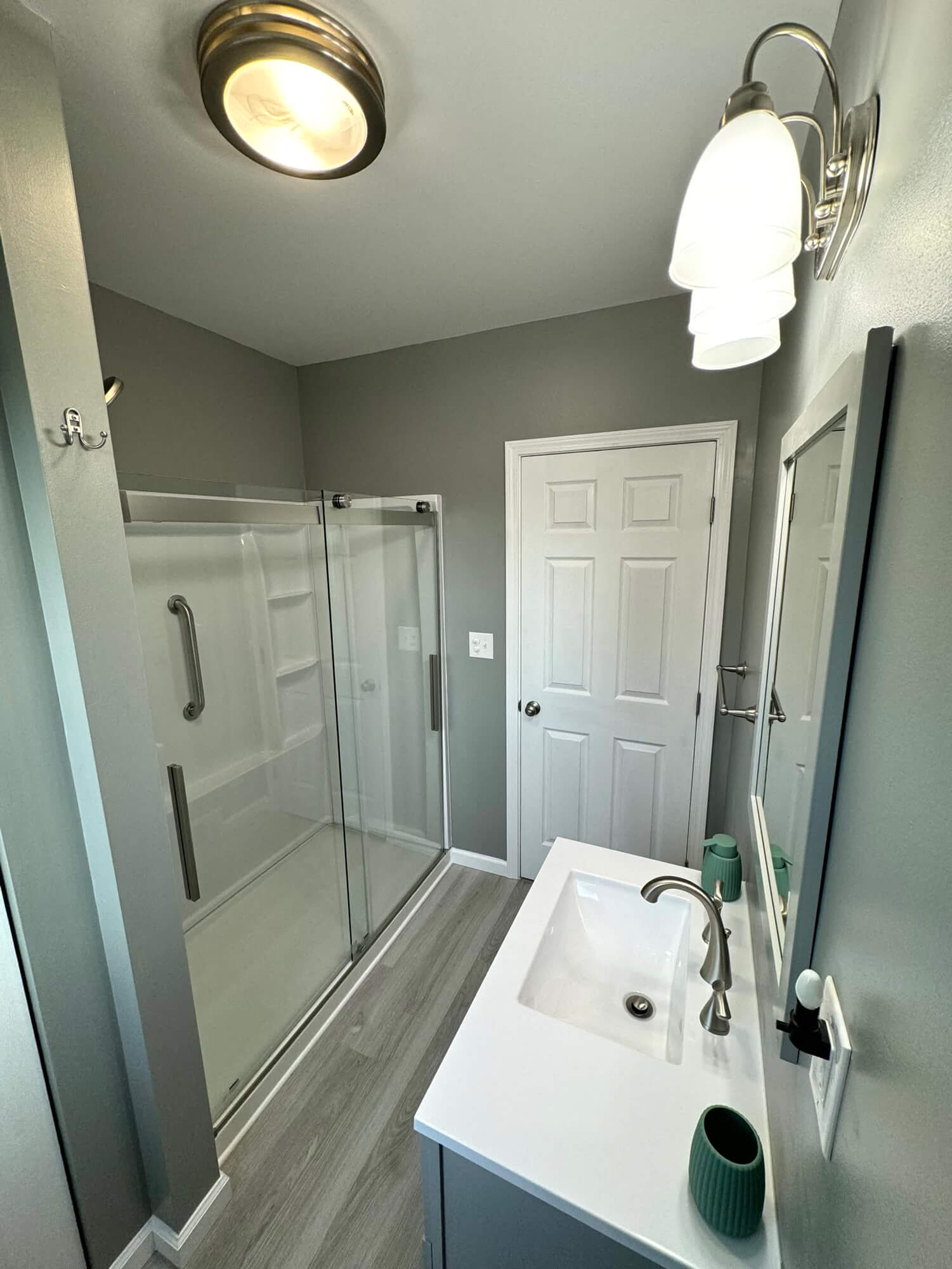 A sleek, newly remodeled bathroom by SC Projects LLC featuring a gray vanity with a white top, modern hardware, a glass-enclosed walk-in shower, and light gray plank flooring.