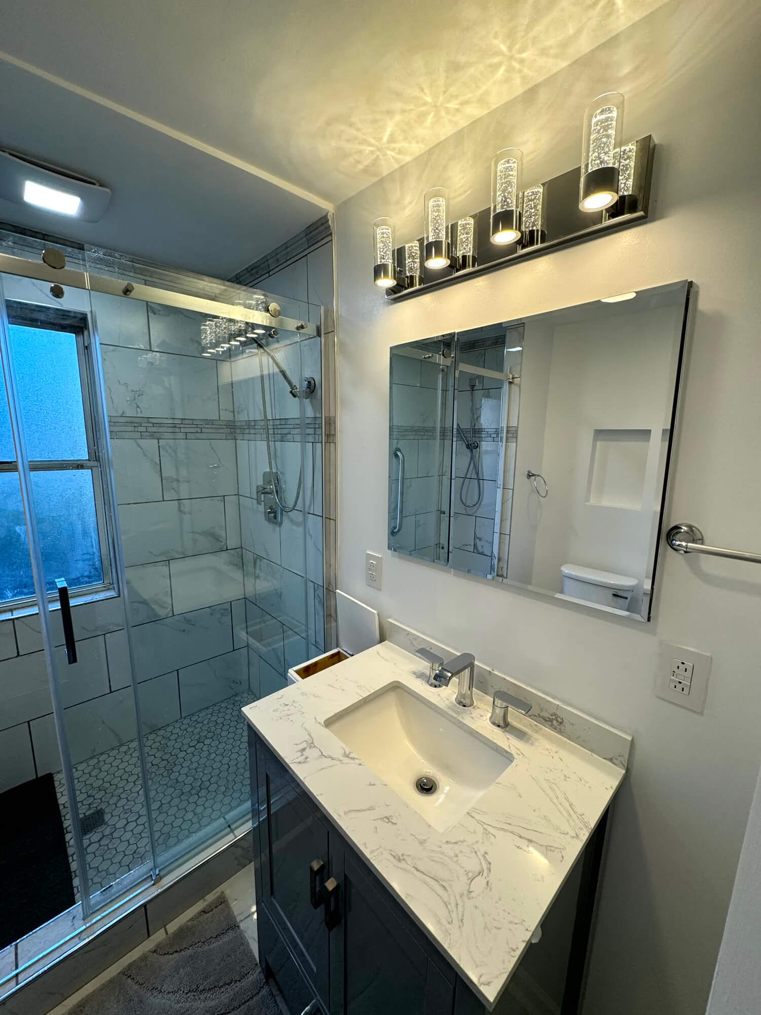 Modern bathroom remodel by SC Projects LLC featuring white marble-look subway shower tile, a glass door, new dark vanity, and contemporary lighting .