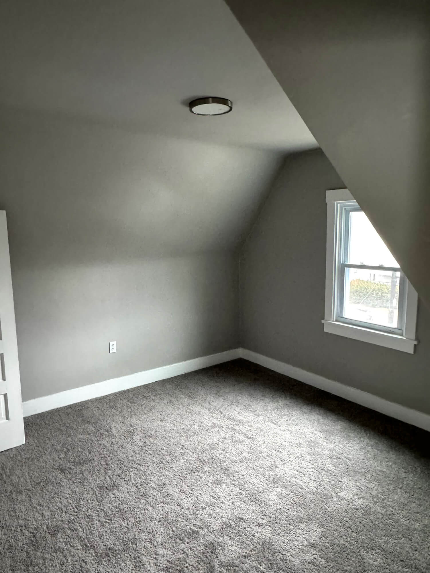 A cozy attic room with gray painted walls, dark gray carpet, and white trim around a small window, showing quality Basement Remodeling in Beaver County, PA .