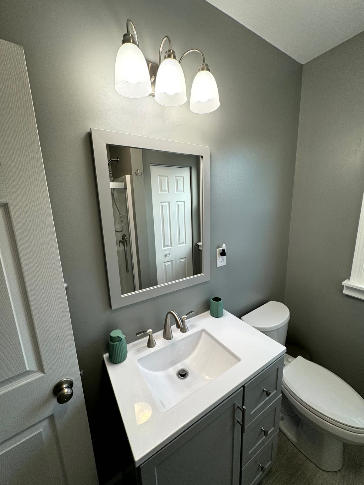 Modern bathroom vanity area with a gray cabinet, white countertop, matching framed mirror, and white toilet against a gray painted wall.