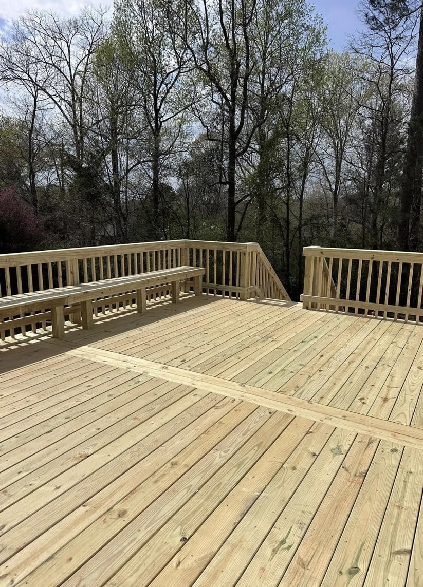 Large new pressure-treated wooden deck construction by SC Projects LLC with built-in bench seating facing a dense forest backyard .
