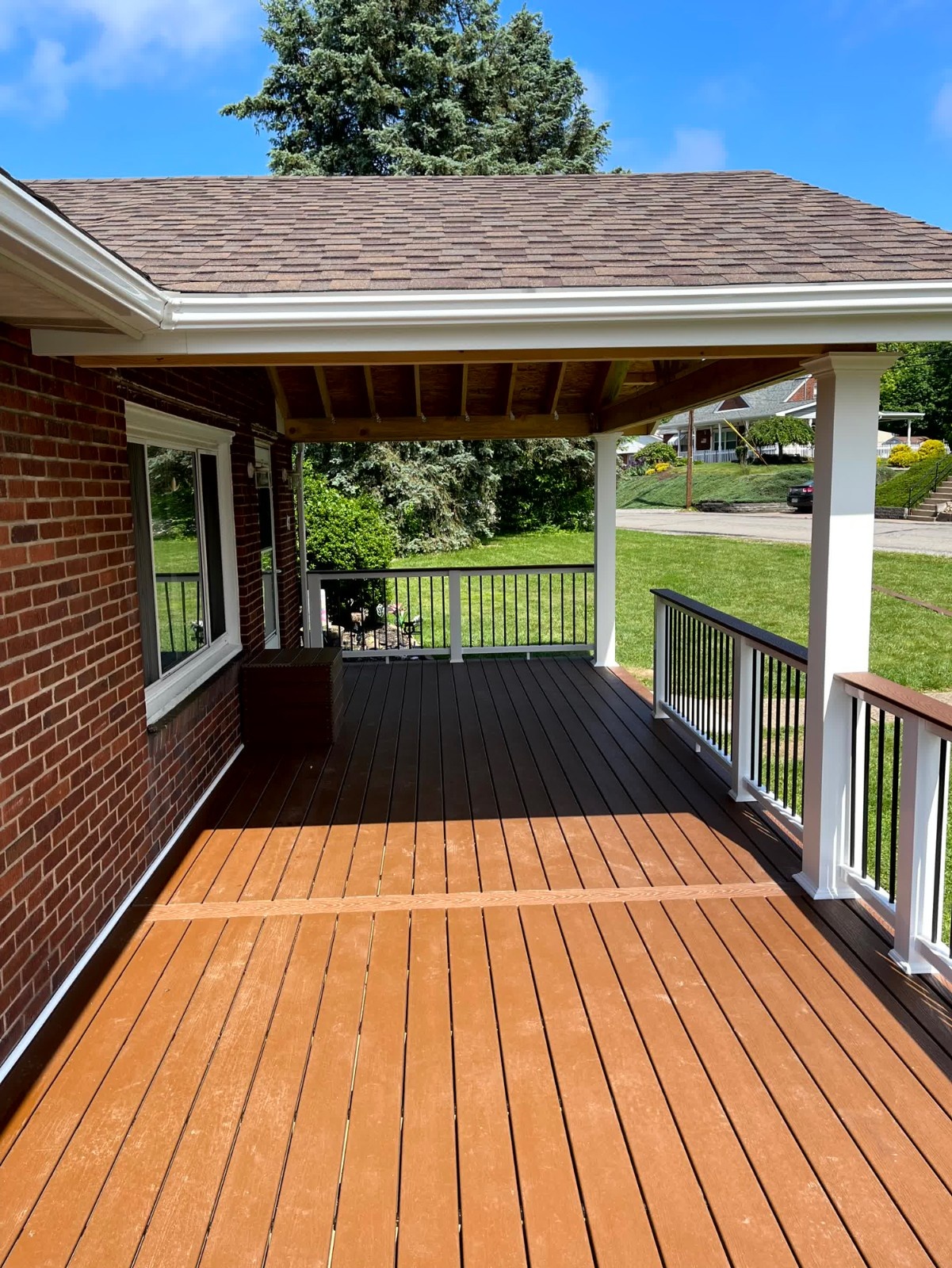 White seamless gutter installed by SC Projects LLC on a covered wood deck attached to a brick home with brown asphalt shingles .