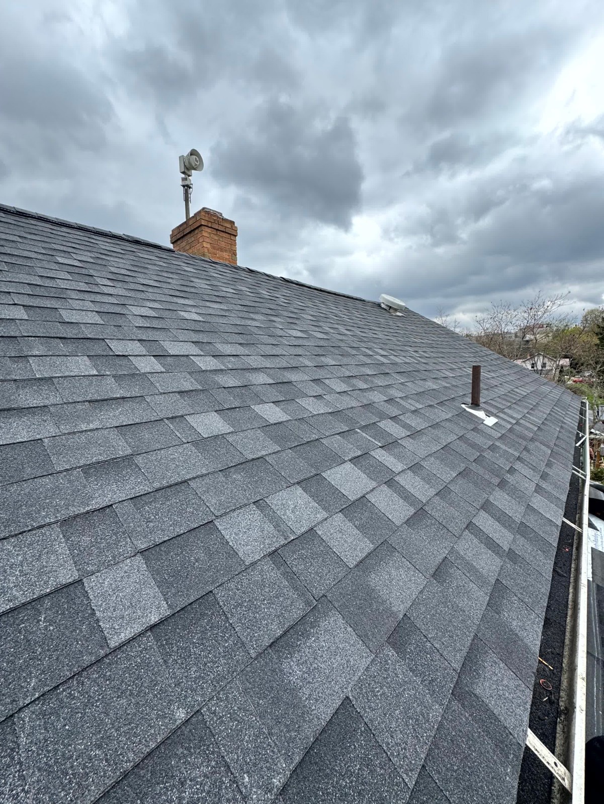 Residential roof replacement with new dark gray asphalt shingles visible under a cloudy sky, with a chimney and plumbing vent stack.