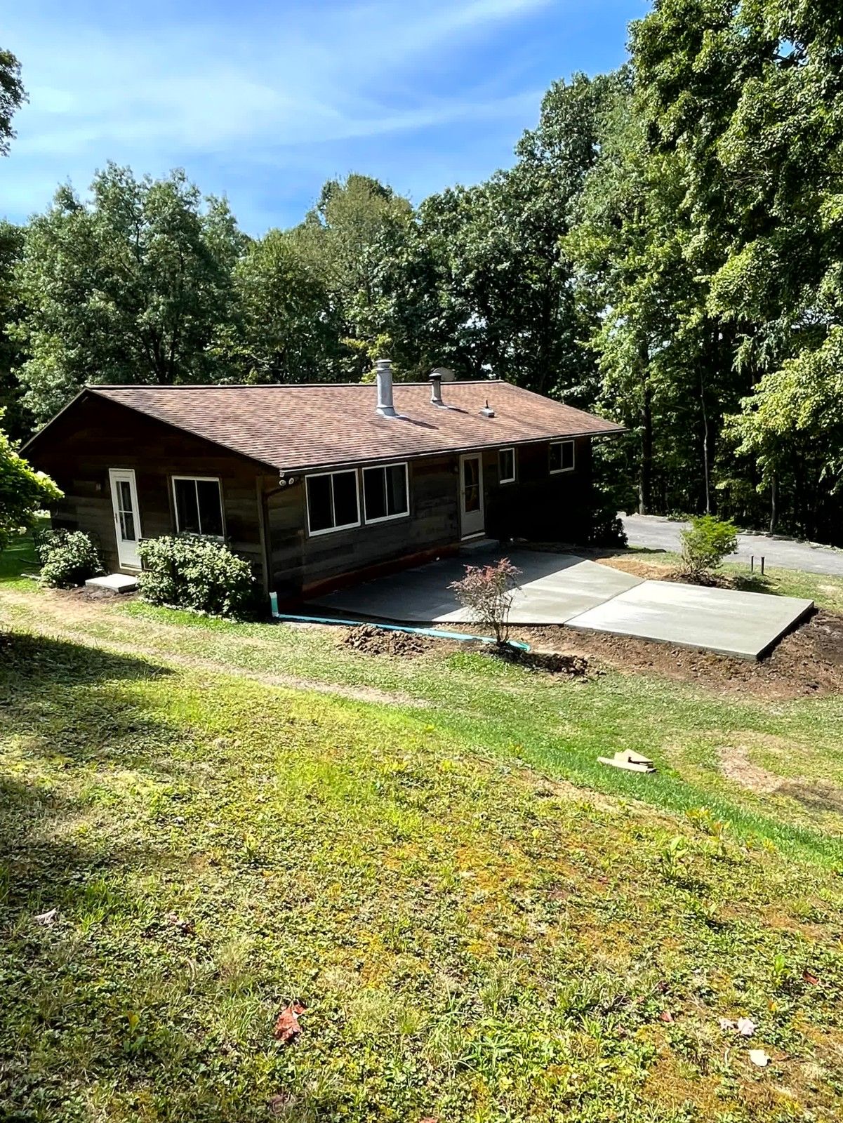 Single-story home with wood siding and a brown shingle roof, featuring a newly poured concrete patio or walkway surrounded by grass and trees.
